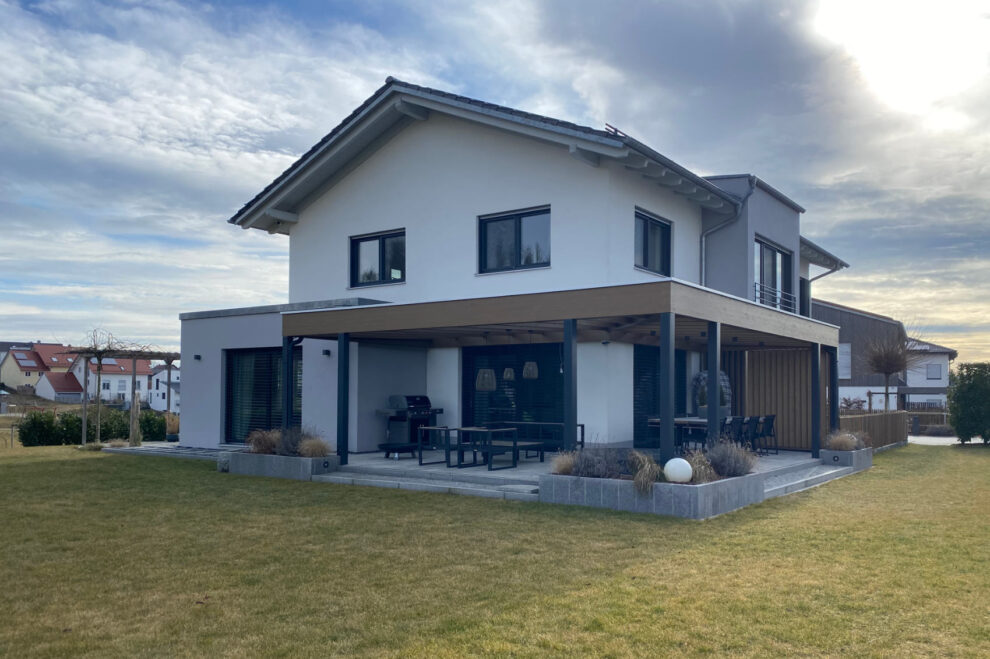Modernes Einfamilienhaus mit weiß-grauer Fassade, großem Garten und überdachter Terrasse mit Sitzbereich und Grill.